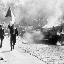 Protestujúci Pražania prechádzajú so štátnou vlajkou popri horiacom okupačnom tanku